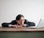 Asian businessman in glasses, resting head on desk, looking at laptop in office setting.